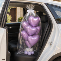 Ramo de globos de corazón lila envuelto en celofán con lazo blanco en el asiento trasero de un auto.