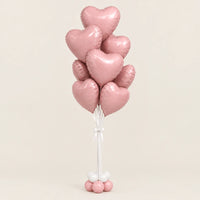 Ramo de globos de corazón rosa metalizado con lazo blanco y base de globitos, sobre fondo beige
